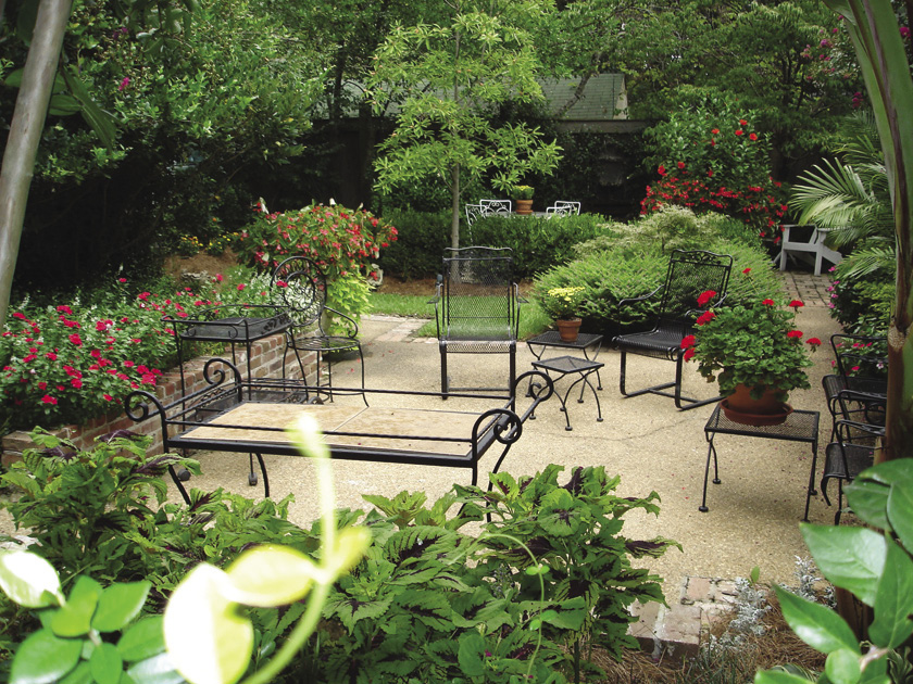 Outdoor dining table and chairs on a concrete patio. Green plants are placed on and around the patio, and small trees and more chairs can be seen farther in the distance.